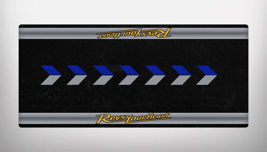 The Motorcycle Pit Mat for Yamaha Universal boasts a black rectangular design with diagonal blue and gray arrows pointing right. Yellow Revs Your Heart text adorns the top and bottom edges on gray bands, evoking the style of a sleek motorcycle floor mat.