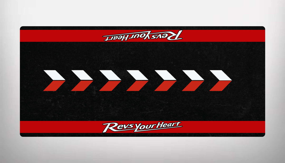 The Motorcycle Pit Mats Yamaha Universal design features a black rectangular mat with red horizontal stripes at the top and bottom. It displays white and red right-pointing arrows in the center and Revs Your Heart text on both stripes, repeated upside down at the top.