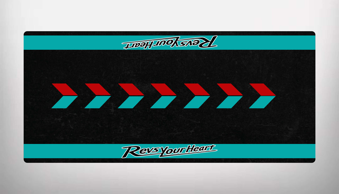 The Motorcycle Pit Mat for Yamaha Universal is a black rectangular mat with red and teal arrows pointing right and features a teal border on top and bottom. Revs Your Heart is elegantly written in white on the borders, perfect for enhancing any motorcycle garage space.