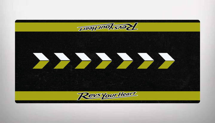 The Motorcycle Pit Mat for Yamaha Universal features a rectangular design with a black base and yellow-green borders. The center displays alternating white and yellow-green arrows pointing right, enhanced by Revs Your Heart text at both the top and bottom for added style.