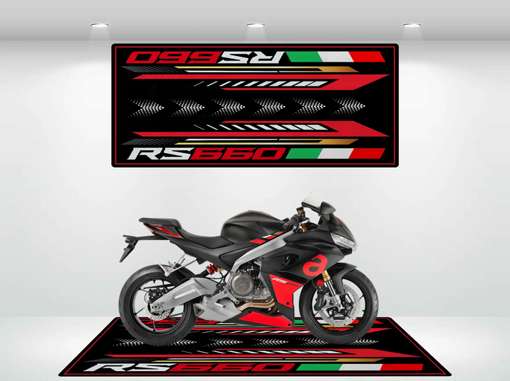 A sleek sportbike is displayed on a black and red Motorcycle Pit Mat, with RS660 and the Italian flag colors prominently featured. The matching backdrop enhances the dynamic allure of the bike in its showroom setting.