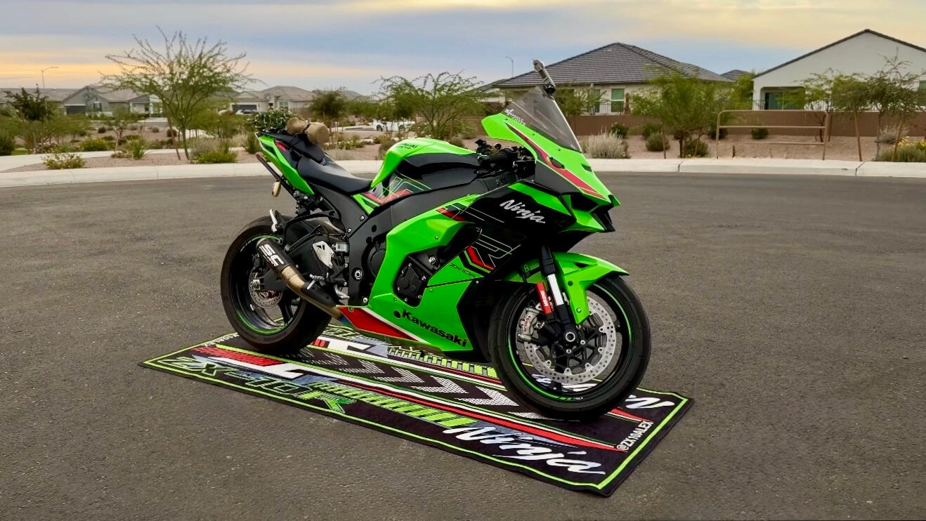 A bright green sport motorcycle with black and red accents is parked on a matching motorcycle mat in an open parking lot. The backdrop features suburban homes, trees, and a cloudy sky.