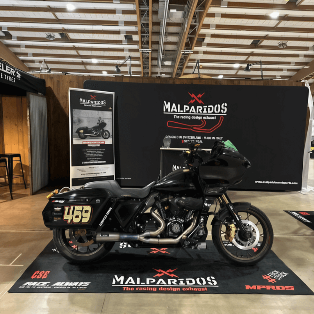 A black motorcycle adorned with the number 469 rests on a platform indoors, complemented by a sleek motorcycle floor mat. The backdrop, showcasing banners for Malparidos, highlights racing design exhausts in an exhibition-like setting.