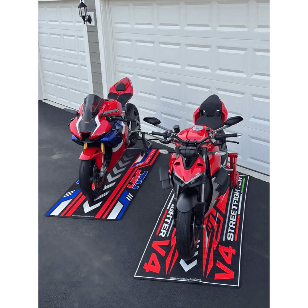 Two red sport motorcycles rest on vibrant motorcycle floor mats in a driveway before white garage doors. One bike boasts fairings, while the other reveals a more exposed design. The mats showcase striking red, blue, and white racing designs, adding flair to the scene.