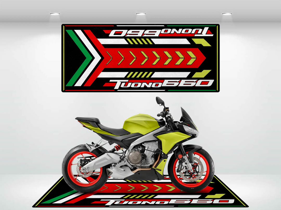 A green and black motorcycle with red wheel rims is showcased in a showroom against a white wall. The motorcycle floor mat, Motorcycle Mat for Tuono 660 (Special Design) by Motorcycle Pit Mat, and a wall banner feature bold arrow patterns in red, white, green, and yellow with Tuono 660 text.