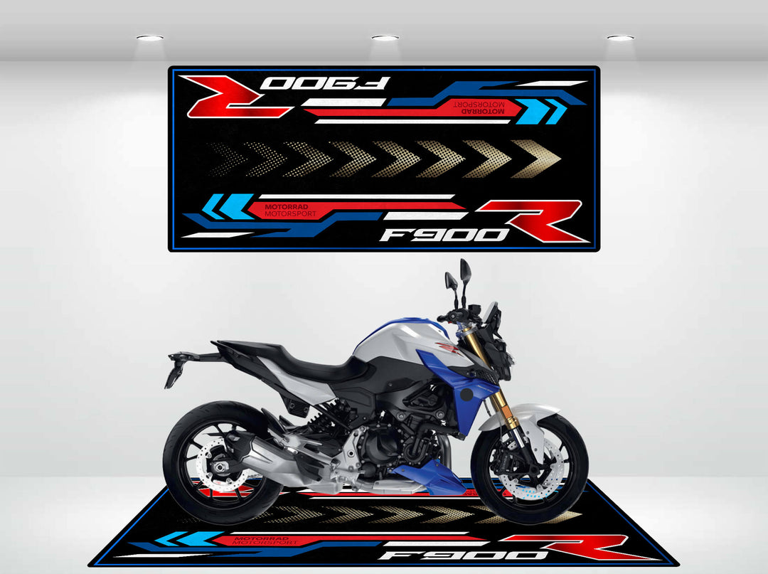 A sporty motorcycle sits on a stylish Motorcycle Pit Mat for F900R, featuring a sleek blue, black, and silver design. Above it hangs a large, colorful F900R banner with dynamic graphics.