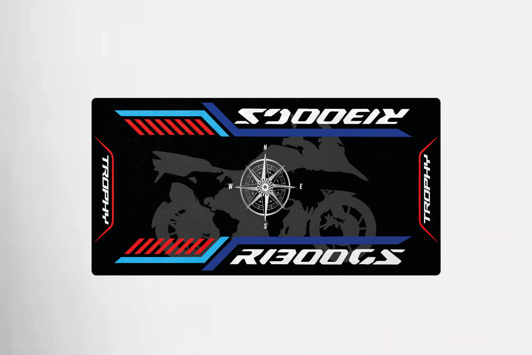 The Motorcycle Pit Mat for R1300 GS Trophy showcases a motorcycle silhouette with a central compass rose on a black background accented by red and blue stripes, featuring R1300GS and Trophy text. An ideal addition to any motorcycle enthusiasts space.