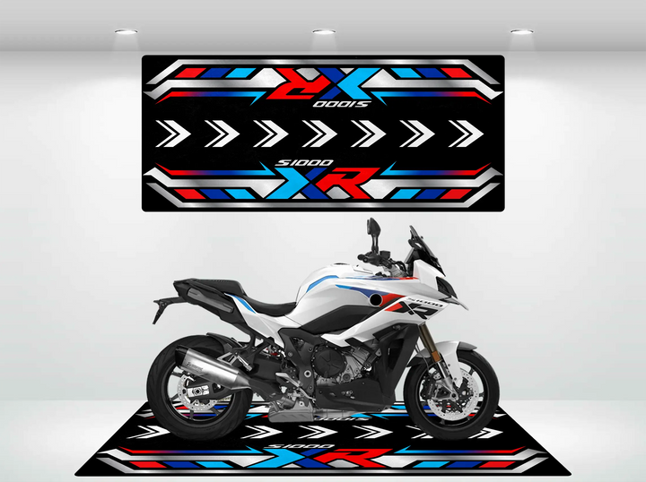 A sleek white motorcycle with blue, red, and black accents is parked on the Motorcycle Pit Mat for the S1000 XR, showcasing geometric designs in matching colors. The backdrop mirrors these patterns with forward-pointing arrows, adding a dynamic touch.