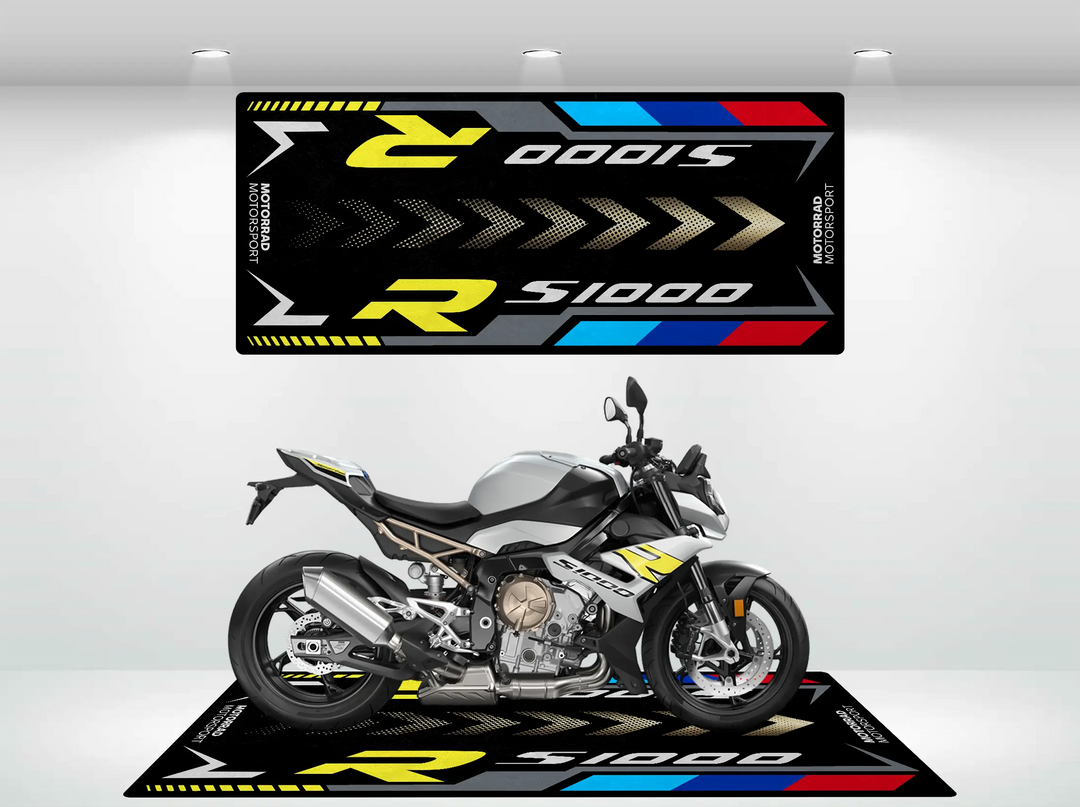 A sleek silver and black motorcycle is displayed on a Motorcycle Pit Mat branded Motorcycle Mat for S1000R, with yellow, white, red, and blue accents featuring S1000 and R logos in a well-lit showroom.