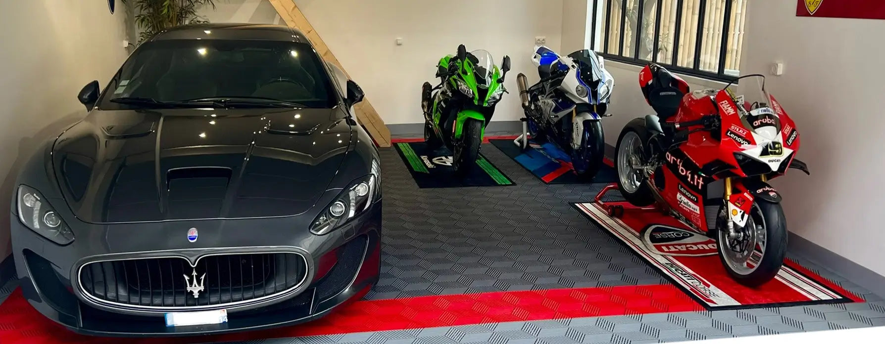 A sleek gray sports car is parked in a garage next to three colorful motorcycles: one green, one blue, and one red. The floor boasts a motorcycle floor mat with an intricate pattern, and a wooden ladder leans against the wall.