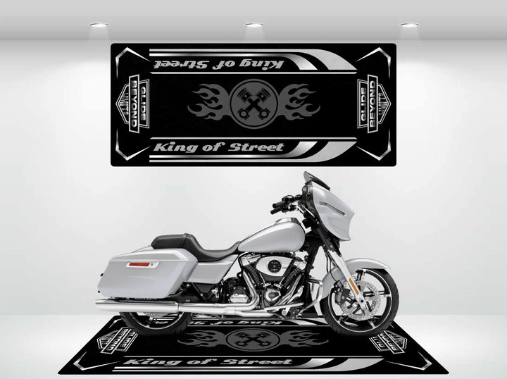 A silver motorcycle rests on the Motorcycle Pit Mats King of Street - Glide Beyond Limits mat, featuring flame patterns and matching banner. Overhead spotlights illuminate the dramatic scene.