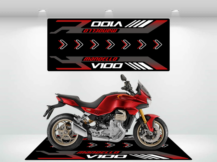 In a showroom, a red V100 Mandello motorcycle stands against a black and red backdrop adorned with white and red arrows. The display features the Motorcycle Pit Mat for Mandello V100 Special, echoing these design elements while highlighting the model name.