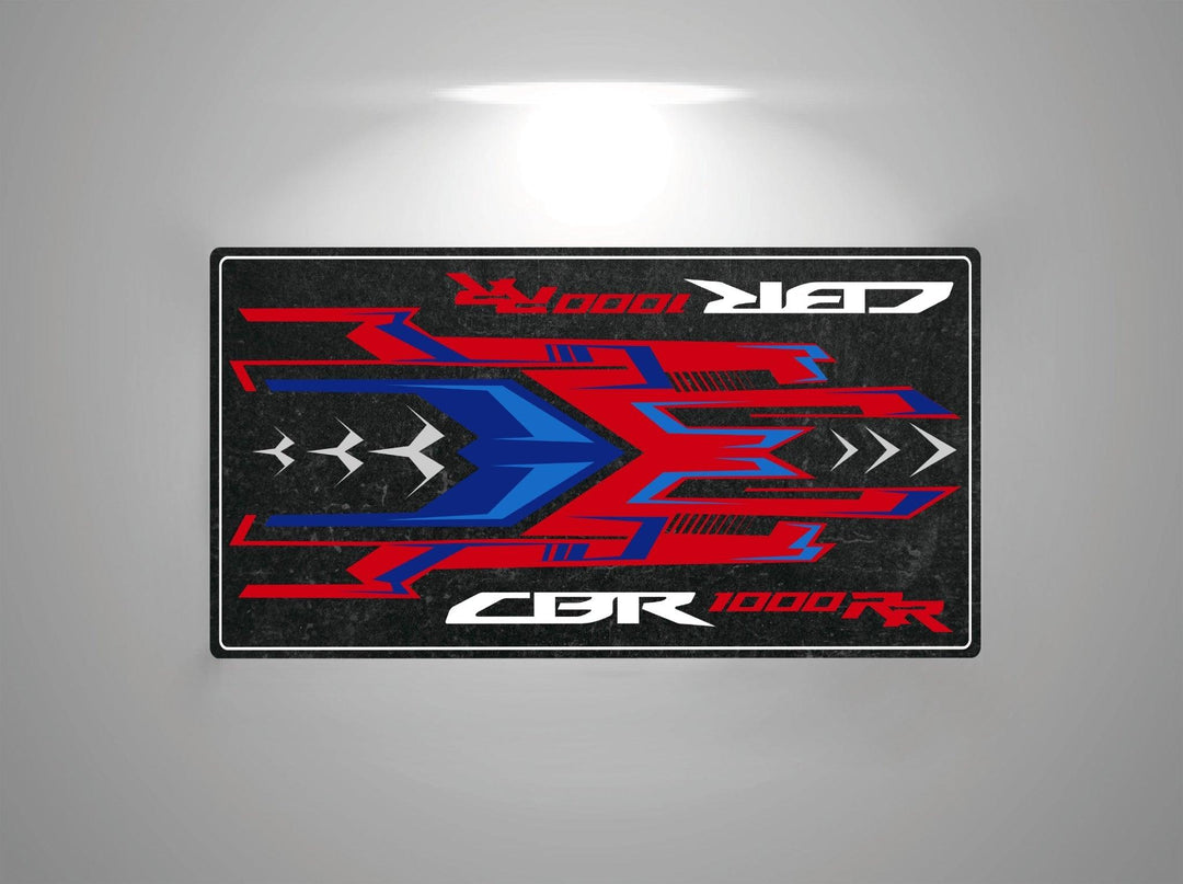 The Motorcycle Pit Mat for Honda CBR1000RR features a rectangular design with abstract red and blue angular shapes on a black background. CBR 1000 RR appears in white text next to mirrored text, enhancing its dynamic and sporty appeal.