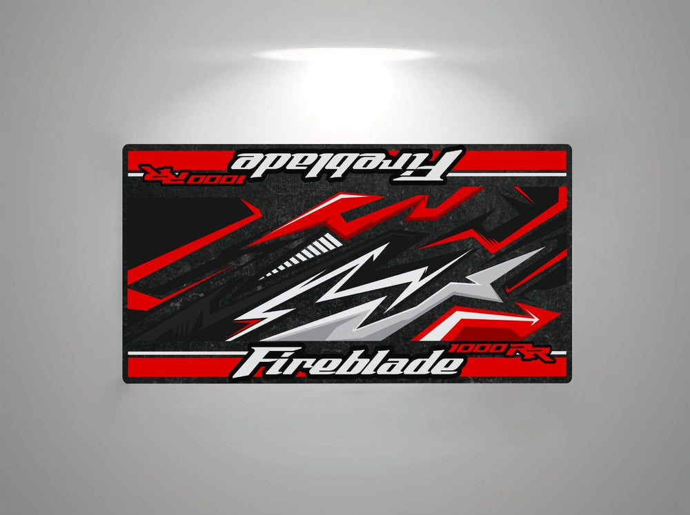 The Motorcycle Pit Mat for Honda CBR1000RR Fireblade is a rectangular mat with an abstract design in black, red, and white, featuring stylized arrows and lightning motifs. It includes Fireblade 1000RR text at both ends—a perfect addition for any motorcycle garage.