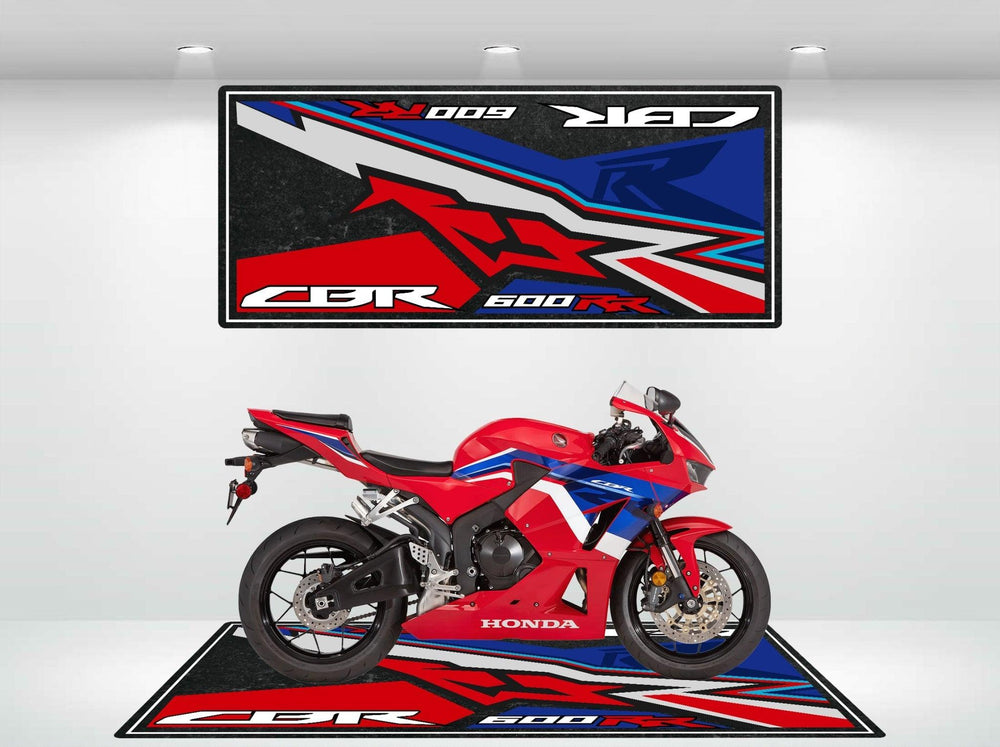 A red and blue Honda CBR600RR motorcycle sits on a Motorcycle Pit Mat, complemented by a large wall hanging with similar red, blue, and black graphics. The room is well-lit with spotlights.