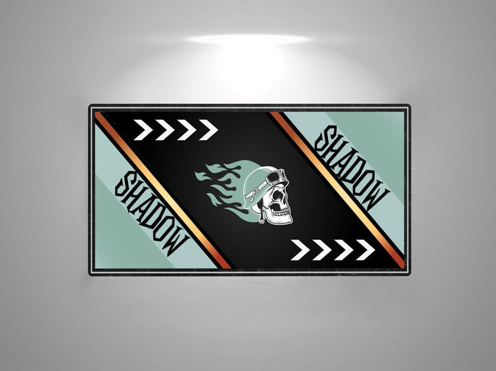 A rectangular image with a black and teal background shows a skull in goggles, embodying the essence of the Motorcycle Mat for Honda Shadow. SHADOW flanks each side with white arrows on teal stripes, illuminated by light above by Motorcycle Pit Mat.