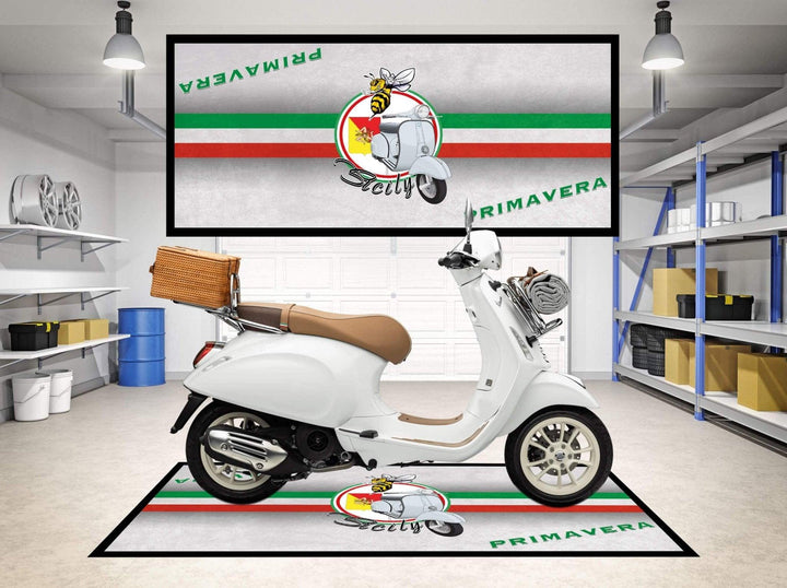 A white scooter with a wicker basket is parked on a stylish Motorcycle Pit Mat for Vespa Primavera in a modern garage. A large banner displaying a bee on a scooter, the word Vespa, and Primavera with Italian flag colors hangs behind it, with shelving units lining the sides.