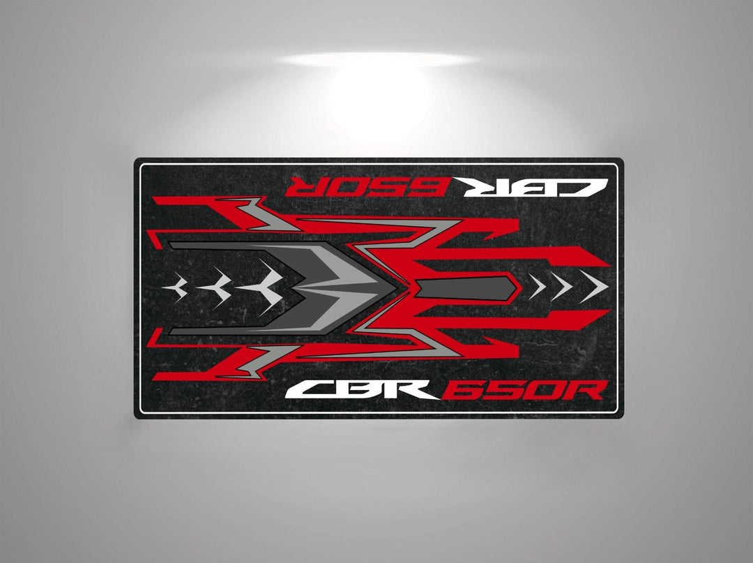 This Motorcycle Pit Mat for Honda CBR650R showcases dynamic red and gray geometric shapes with arrows, alongside the text CBR 650R on top and bottom. Set against a rugged dark backdrop resembling a garage mat, it features a spotlight effect illuminating the design.