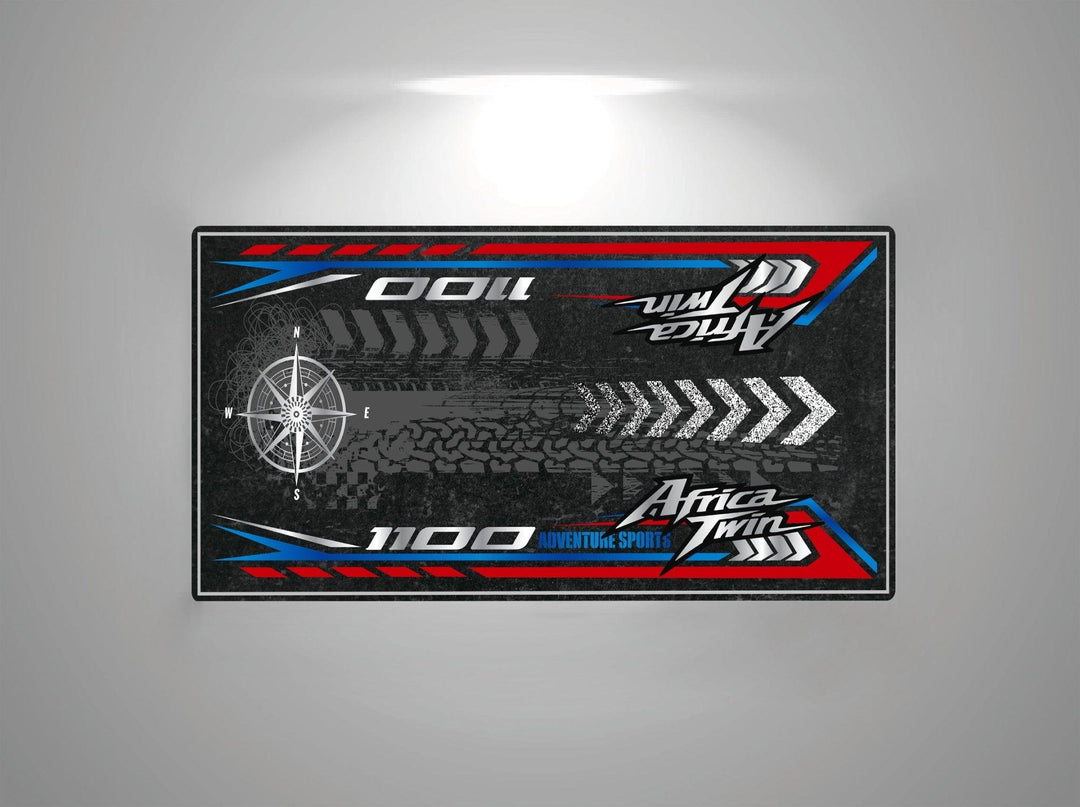 The Motorcycle Pit Mat for Honda Africa Twin features a sleek design with a black background, red and blue accents, tire tracks, compass, arrows pointing right, and the text Africa Twin 1100 and Adventure Sports, perfect for any passionate riders space.