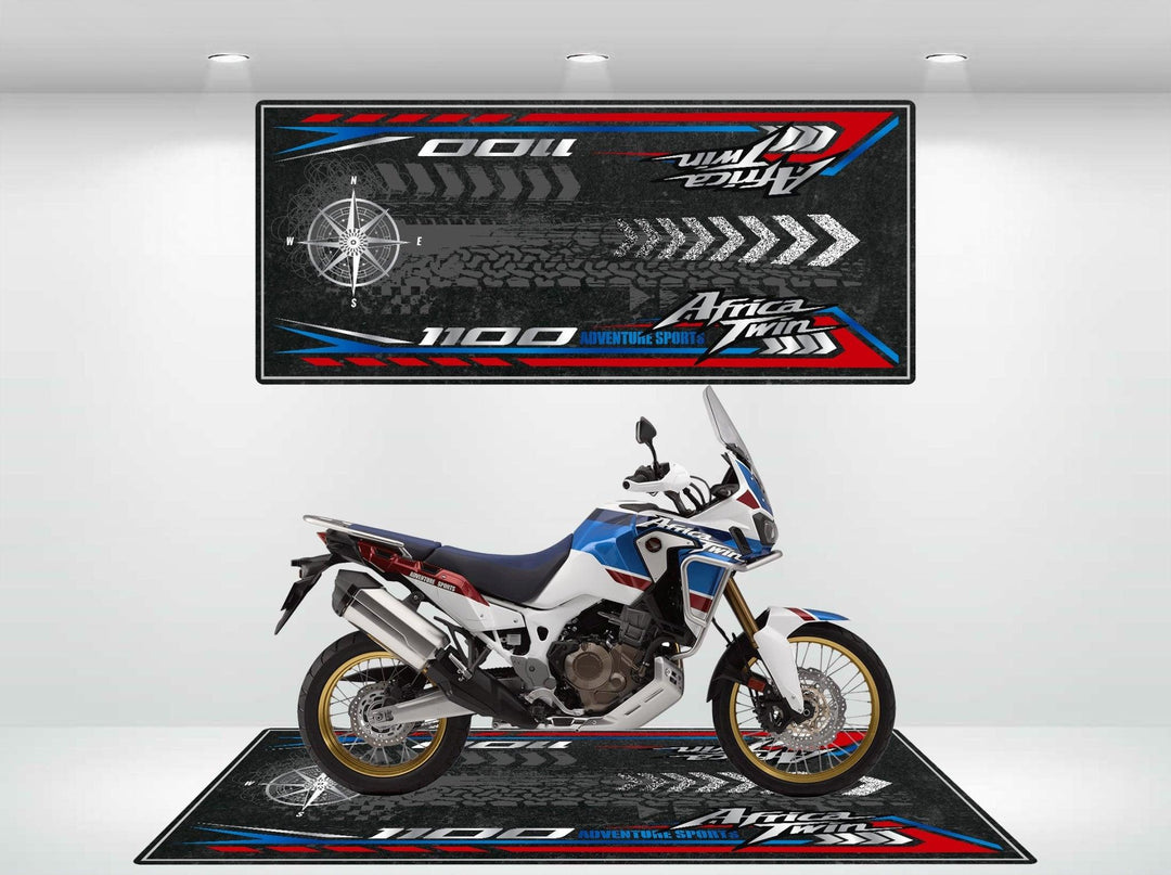 A Honda Africa Twin motorcycle rests on a vibrant Motorcycle Pit Mat in the showroom. The bike is red, white, and blue with golden rims. The mat features tire tracks, a compass, and 1100 Africa Twin Adventure Sports, echoing the adventurous spirit of the display.