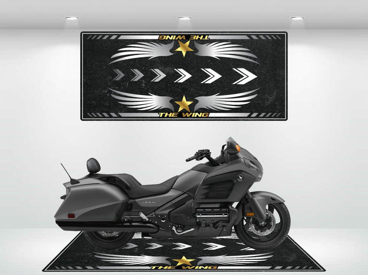 A black motorcycle is parked on a Motorcycle Pit Mat for Honda Goldwing, featuring a bold design with wing motifs, stars, and THE WING in yellow. A matching graphic enhances the wall behind it.