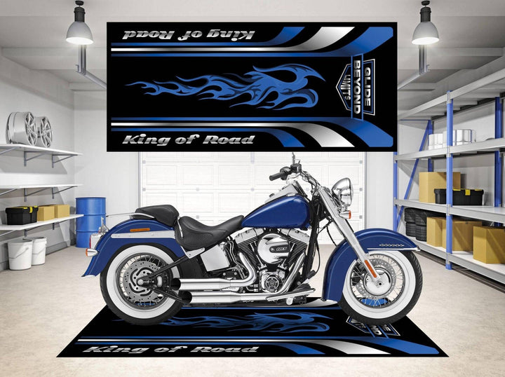 A blue motorcycle rests on a Motorcycle Pit Mat, specifically the Harley King of Road model, inside a garage. Shelves with storage bins and car-related items line the walls, while a matching flame wall banner enhances the decor.