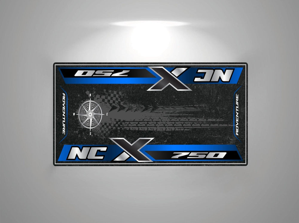 A rectangular motorcycle mat from Motorcycle Pit Mat, designed for the Honda NC750X, features NCX 750 and Adventure with a compass graphic, all in blue over a dark textured background.