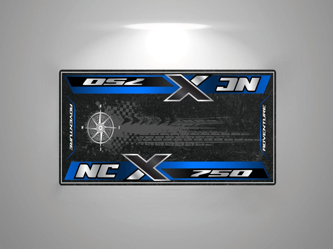 A rectangular motorcycle mat from Motorcycle Pit Mat, designed for the Honda NC750X, features NCX 750 and Adventure with a compass graphic, all in blue over a dark textured background.