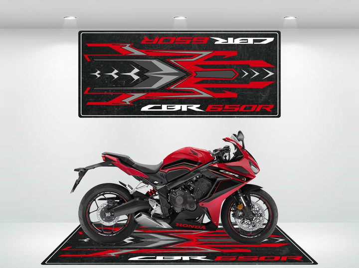 A Honda CBR650R in red and black is parked on a Motorcycle Pit Mat for Honda CBR650R, set against a white background. A similar mat hangs on the wall, highlighting the bikes design elements.