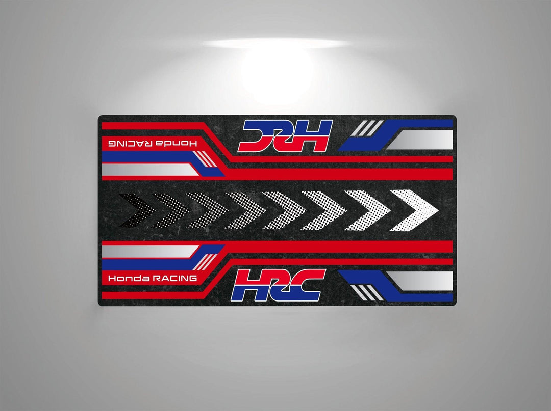 The Motorcycle Pit Mat for Honda HRC features a rectangular design with bold racing-inspired geometric patterns in red, blue, black, and white, showcasing Honda Racing and HRC prominently. It is illuminated from above to highlight its vibrant colors.