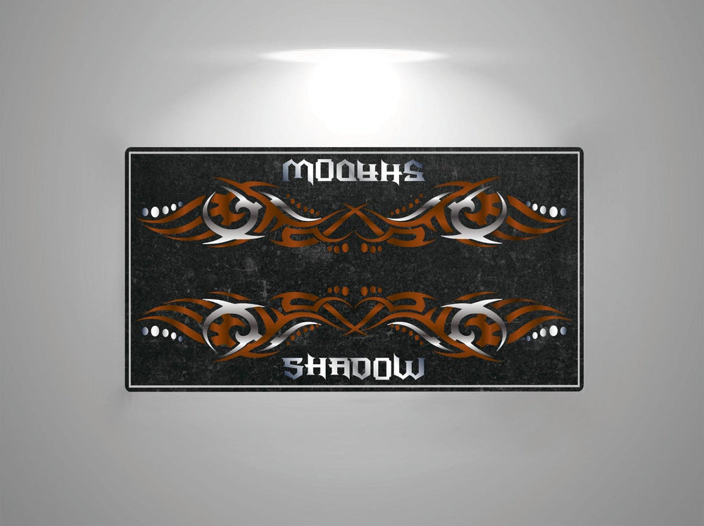 A dimly lit gray motorcycle mat, branded as Motorcycle Pit Mat, showcases a black rectangular design with intricate brown and white tribal patterns. The words Honda Shadow Brown adorn the top and bottom, separated by the designs.