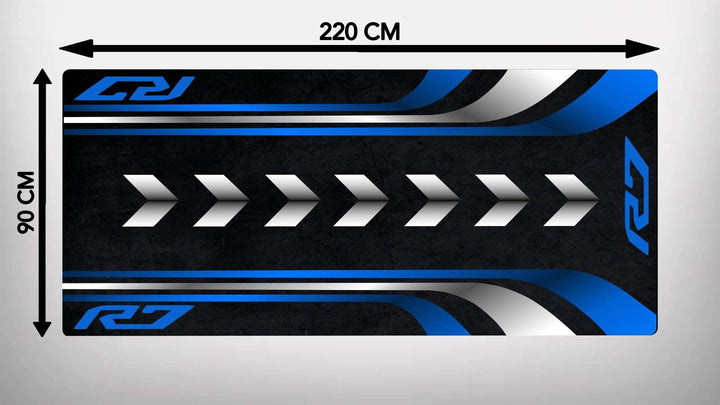 The Motorcycle Pit Mat for Yamaha R7 is a 220 cm by 90 cm rectangular garage mat with a black background, blue and white curved side stripes, and central white arrows pointing right. Stylized R logos adorn the corners, making it ideal for any motorbike enthusiasts space.