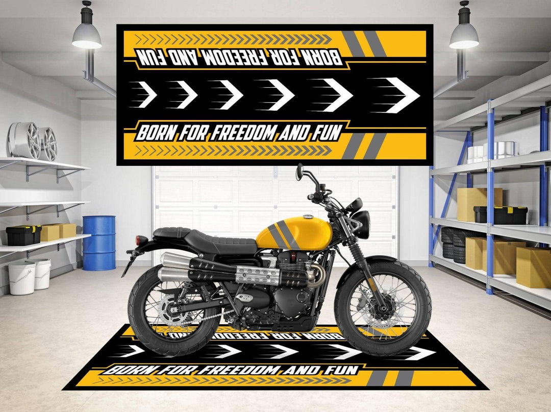 A yellow motorcycle rests on a Motorcycle Pit Mat for Triumph Scrambler in a garage, which is adorned with black and yellow Born for Freedom and Fun banners. Shelving and various garage items lie scattered in the background.