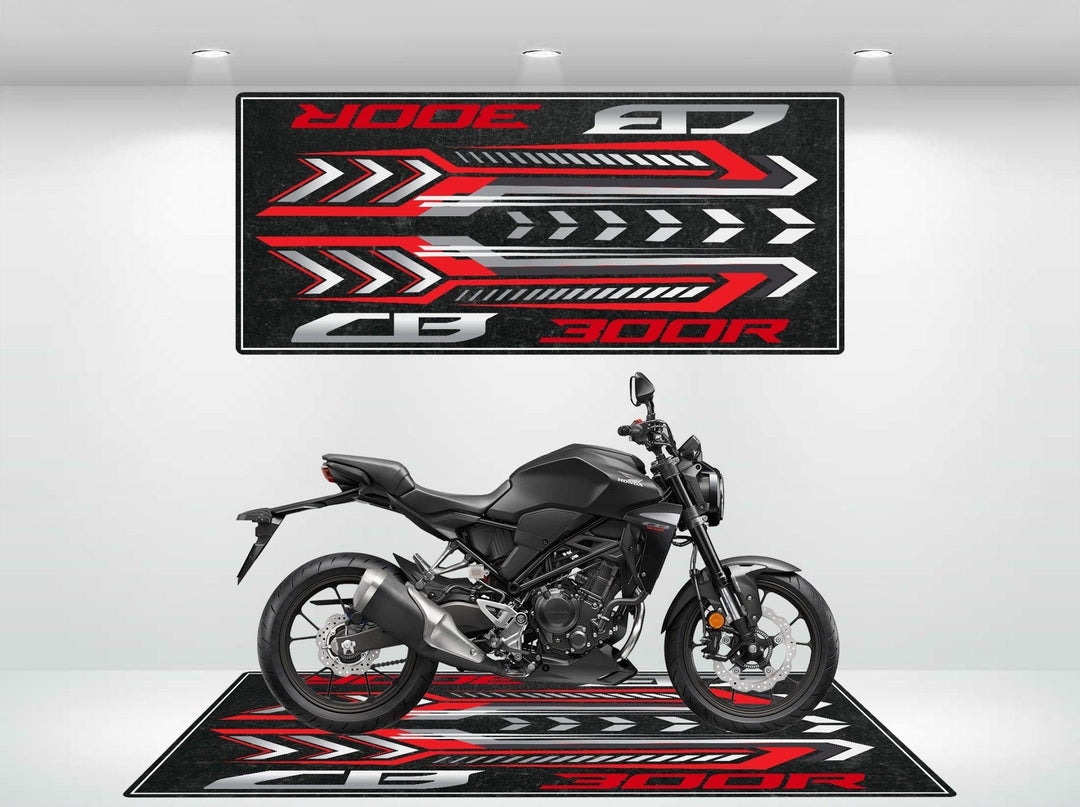 A sleek black motorcycle is parked indoors on a Motorcycle Pit Mat sporting bold red, black, and white graphics. Above it, the same design features the Honda CB300R text. Bright lighting highlights this setup.