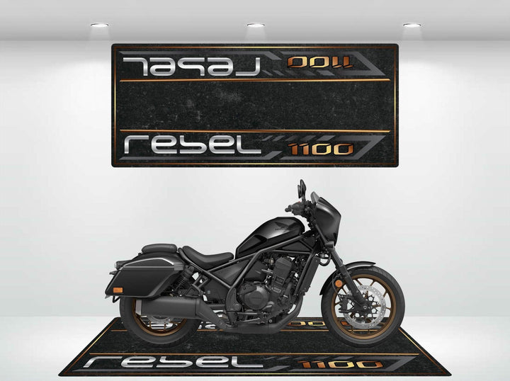 A black motorcycle sits on a Motorcycle Pit Mat designed for the Honda Rebel 1100, under a spotlight. The wall and floor showcase logos and Rebel 1100 in stylized text, creating a modern showroom ambiance that highlights the bikes sleek design.