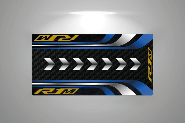 The Motorcycle Pit Mat X JDL MOTO Yamaha R1M Mat displays a rectangular design with white arrows on a carbon fiber background, framed by blue, yellow, and metallic silver accents. Stylized RM text appears on the top and bottom edges for a garage mat flair.