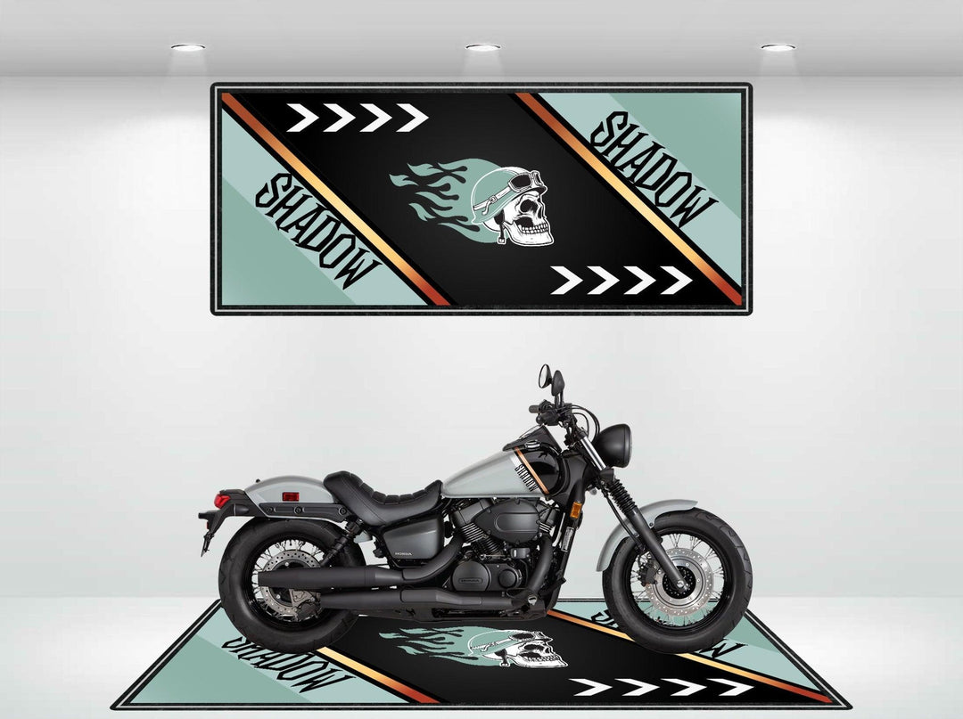 A motorcycle is parked on a Motorcycle Pit Mat for Honda Shadow with matching wall art above, featuring a stylized skull design with flames and the word SHADOW in bold. The color scheme includes black, light blue, orange, and white.