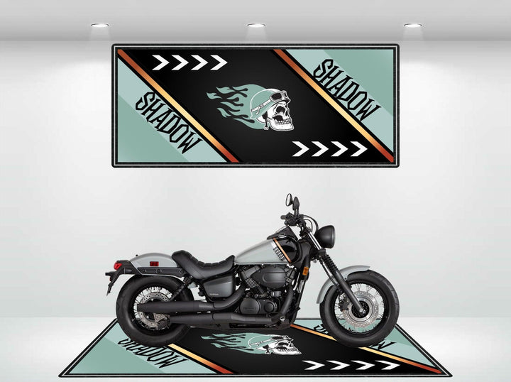 A motorcycle is parked on a Motorcycle Pit Mat for Honda Shadow with matching wall art above, featuring a stylized skull design with flames and the word SHADOW in bold. The color scheme includes black, light blue, orange, and white.