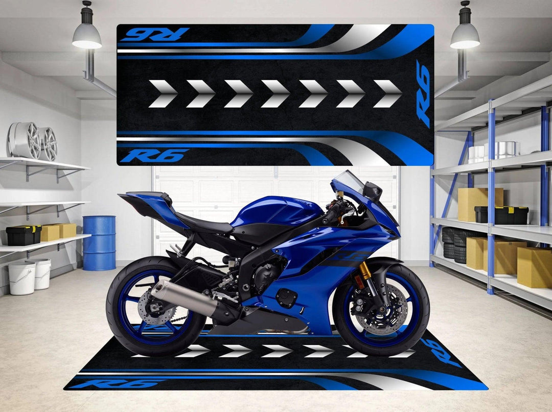 A Yamaha R6 sits on a Motorcycle Pit Mat in a garage with blue and black R6 arrow designs on the floor and wall. The space is clean and organized, surrounded by shelves and items, under overhead lighting.