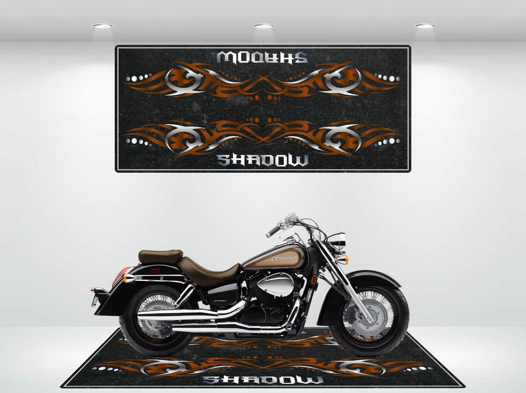 A black motorcycle sits in a showroom on the Motorcycle Pit Mat for Honda Shadow Brown. The mat features tribal-style orange patterns with Shadow in white. A matching wall design creates harmony, while bright overhead lights illuminate the scene.