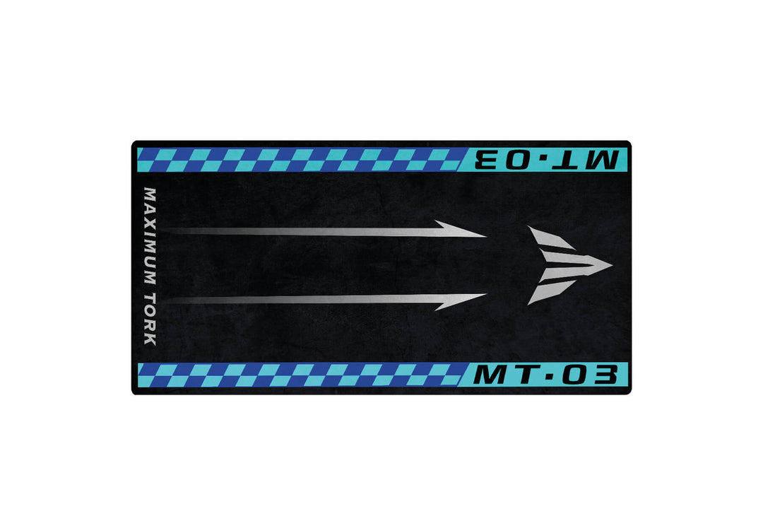 A rectangular Motorcycle Pit Mat for Yamaha MT-03 displays silver right-facing arrows against a black background. Blue and turquoise checkered borders line the top and bottom, with MAXIMUM TORK on the left and MT-03 and E0.LW on the right.