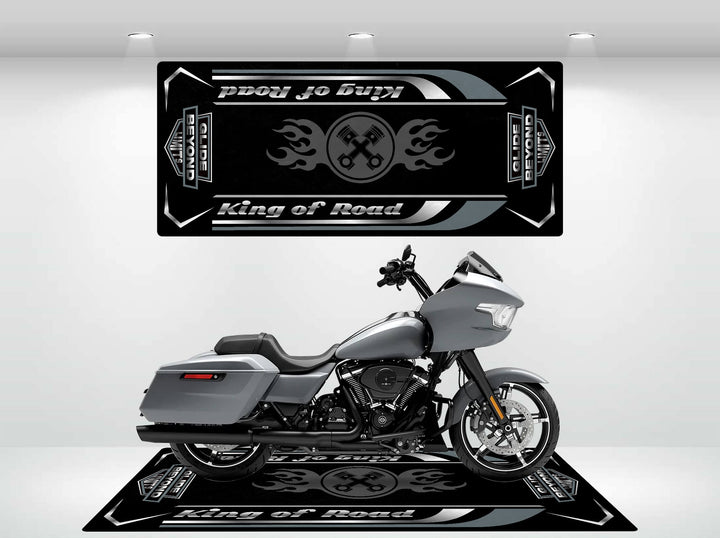 A sleek gray motorcycle is showcased on a black platform with crossed wrenches and flames, set against a King of Road backdrop. The Motorcycle Pit Mat - Glide Beyond Limits enhances the display, ensuring every detail stands out in the well-lit setting.