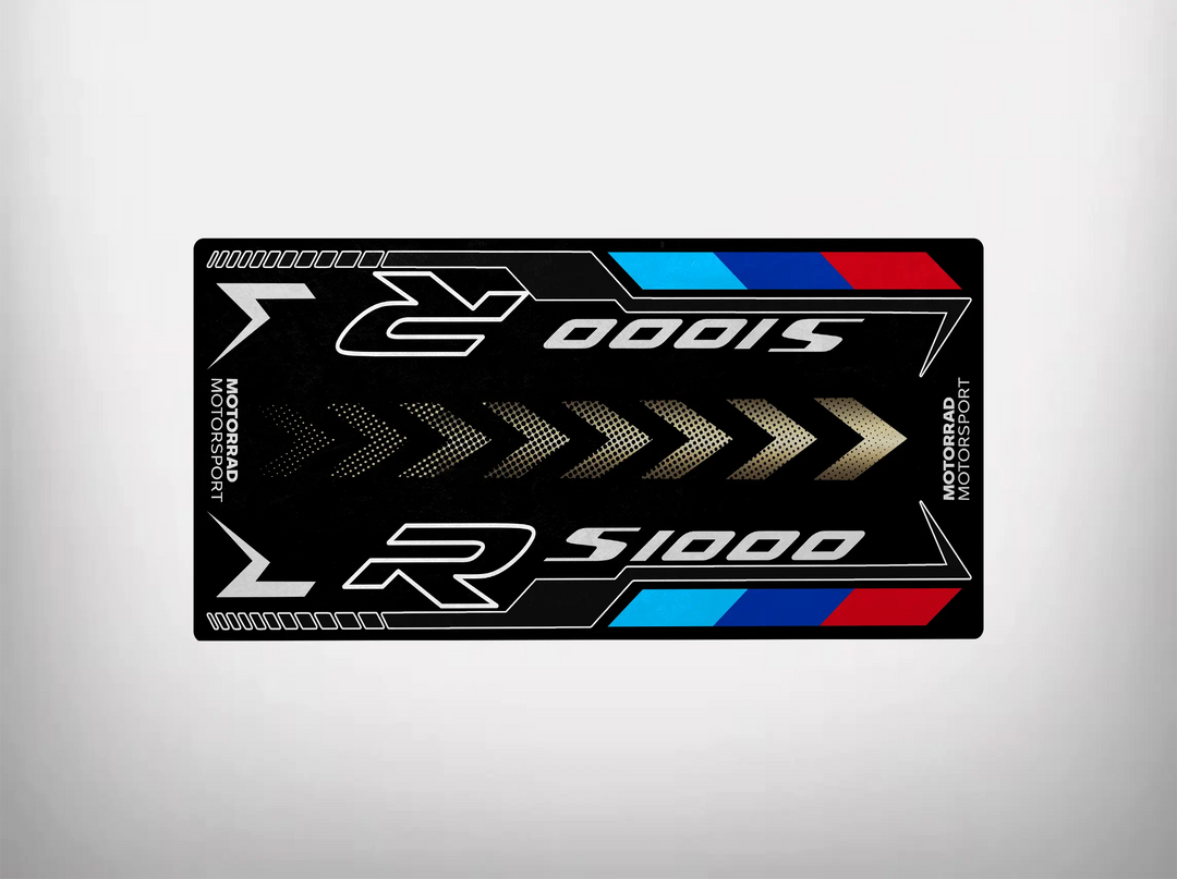 The Motorcycle Pit Mat for S1000R is a black, rectangular sticker with S1000 R in metallic letters and red, blue, and white stripes. It features Motorsport vertically on the edges with arrows pointing inward.