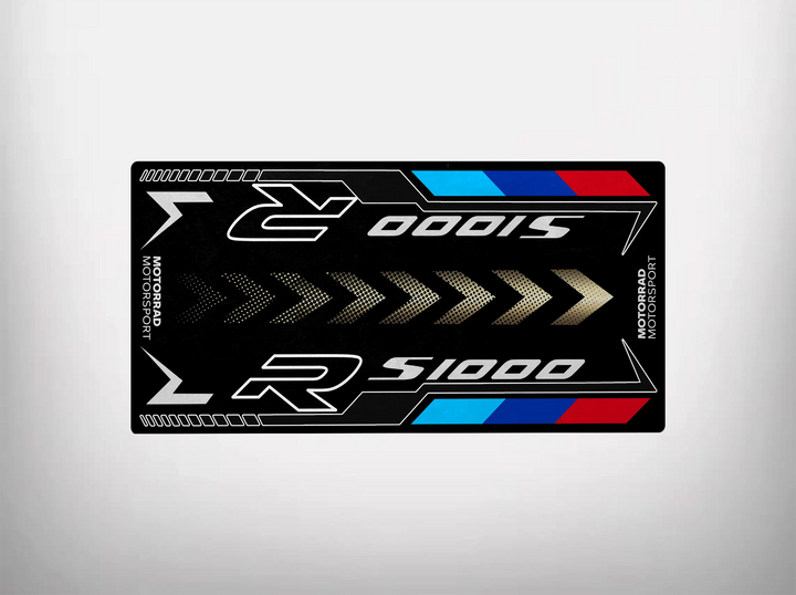 The Motorcycle Pit Mat for S1000R is a black, rectangular sticker with S1000 R in metallic letters and red, blue, and white stripes. It features Motorsport vertically on the edges with arrows pointing inward.