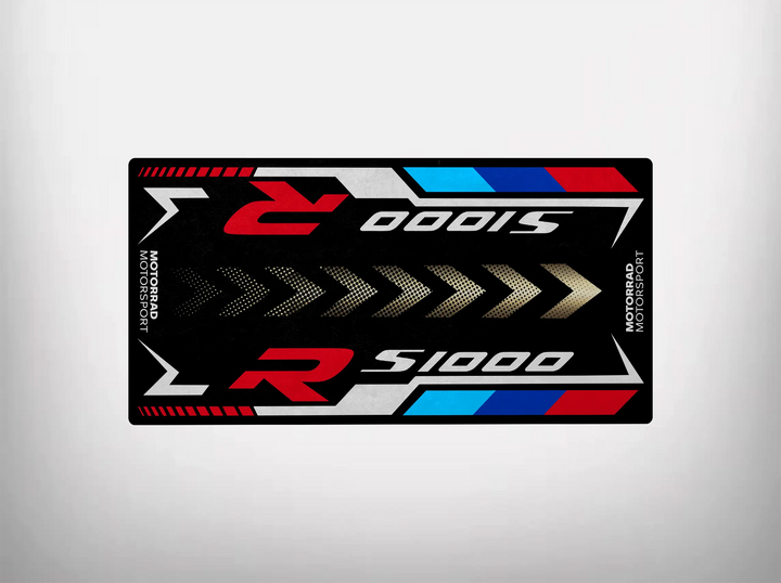 The Motorcycle Pit Mat for the S1000R features a rectangular racing sticker design with R S1000, incorporating red, black, white, and blue arrow patterns. Its modern style enhances garage aesthetics with Motorsport Motorrad on both sides, complementing any motorcycle space perfectly.