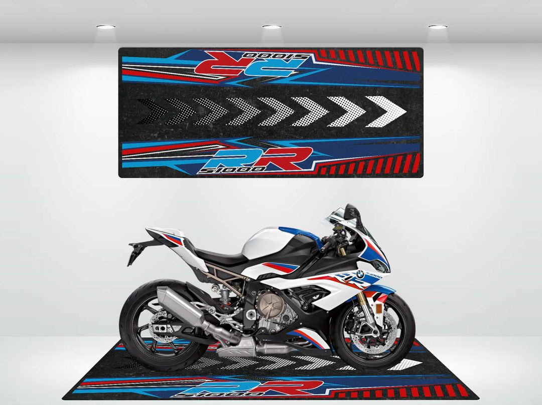 A sport motorcycle in white, blue, and red is parked on a Motorcycle Pit Mat designed for the S1000RR. A matching large panel with bold graphics and arrows is displayed on the wall. The room is brightly lit.