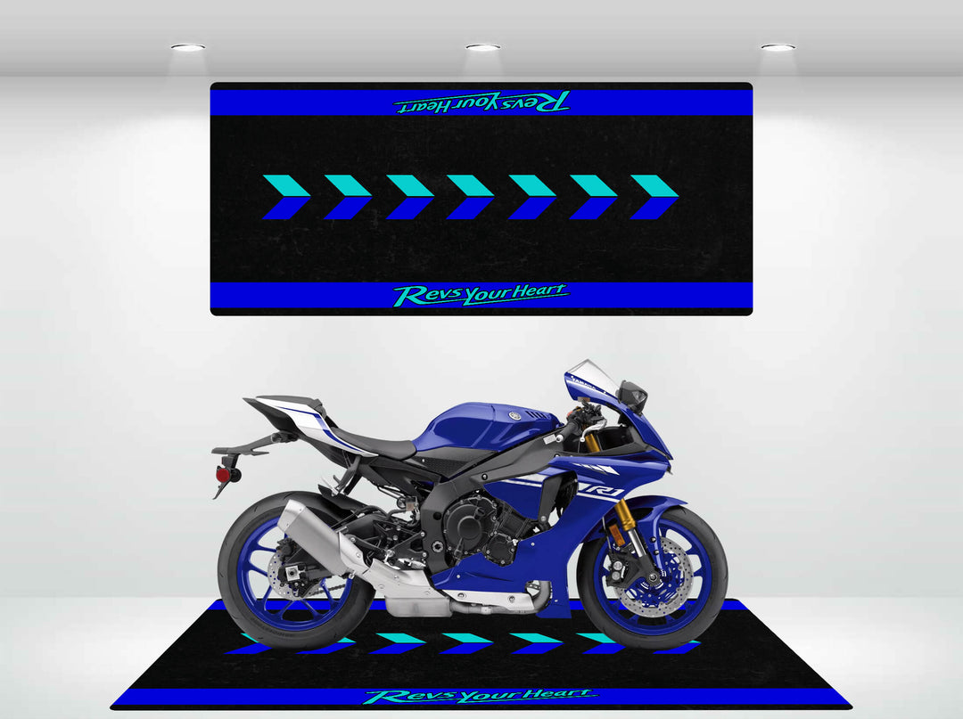 A sleek, blue sport motorcycle sits on a platform against a black and blue backdrop. The Motorcycle Pit Mat for Yamaha Universal adds style with arrow designs and Revs Your Heart in green. Bright overhead lights enhance the display.