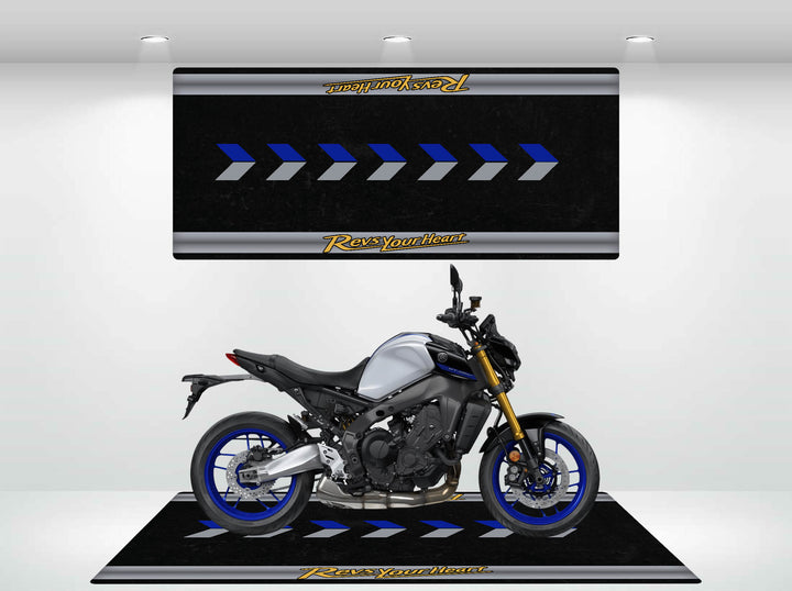 A sleek silver and blue motorcycle rests on a stylish Motorcycle Pit Mat for Yamaha Universal indoors, illuminated by overhead spotlights. The backdrop features a pattern of blue arrows with the bold phrase Rev Your Heart adorning the top and bottom.