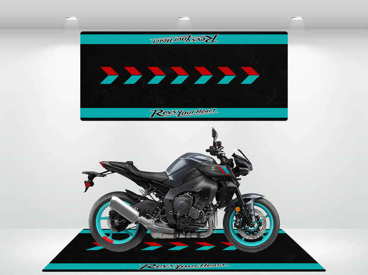 A sleek black motorcycle with turquoise rims sits in a showroom on the Yamaha Universal Motorcycle Mat by Motorcycle Pit Mat. The backdrop and mat feature diagonal arrows and Revs Your Heart in turquoise and red accents, all illuminated by bright overhead lights.