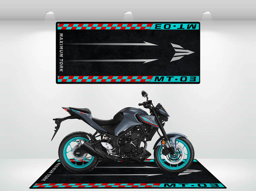 In a showroom, a black and gray motorcycle with turquoise accents stands on a Motorcycle Pit Mat for Yamaha MT-03, featuring MT-03 and Maximum Torque text along with arrows and checkered patterns to enhance the bike’s presentation.